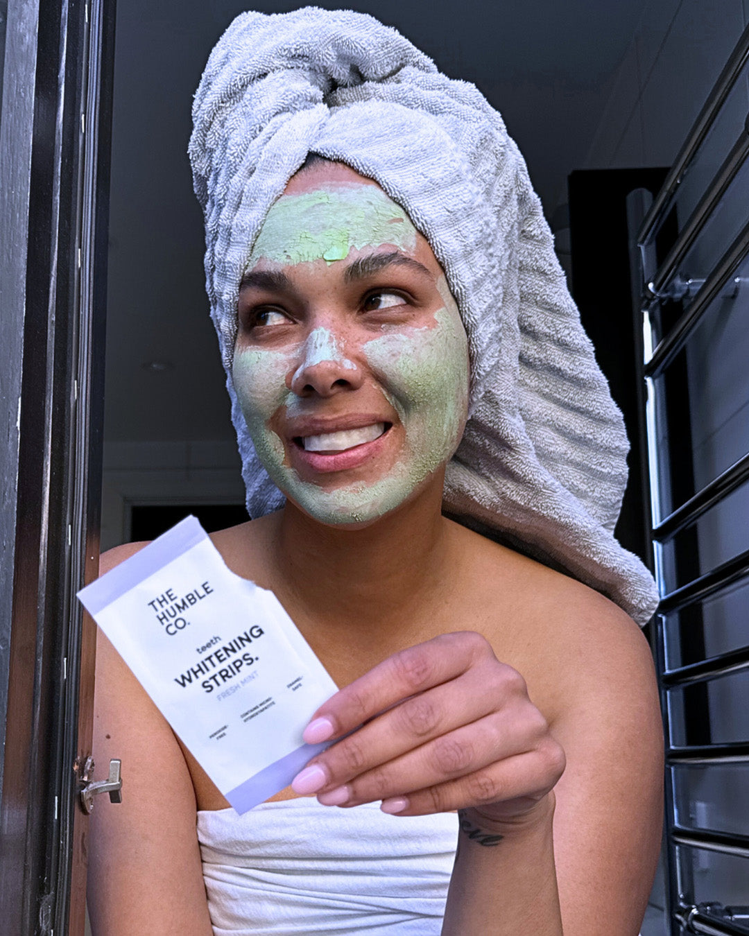 Woman with a green face mask holding Whitening Strips packaging, wrapped in a towel.