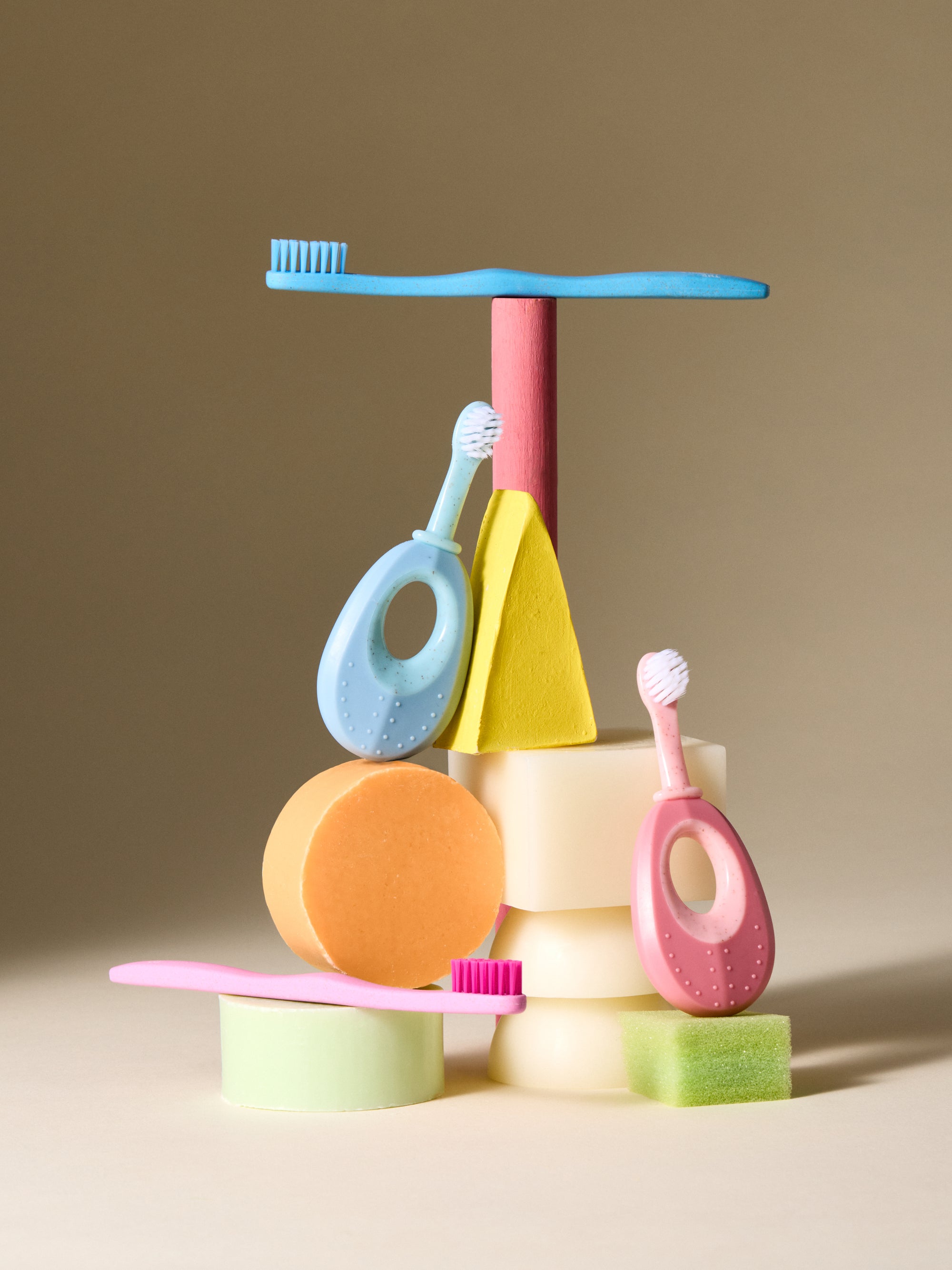 A collection of colorful toothbrushes and dental hygiene products including a blue toothbrush on top of a pink and yellow toothbrush holder, with a pink and yellow sponge below. The products are arranged against a neutral background.