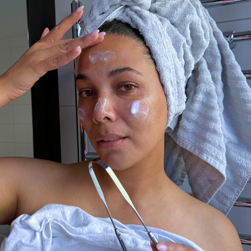 Woman applying cream to her face with a towel on her head in a bathroom setting