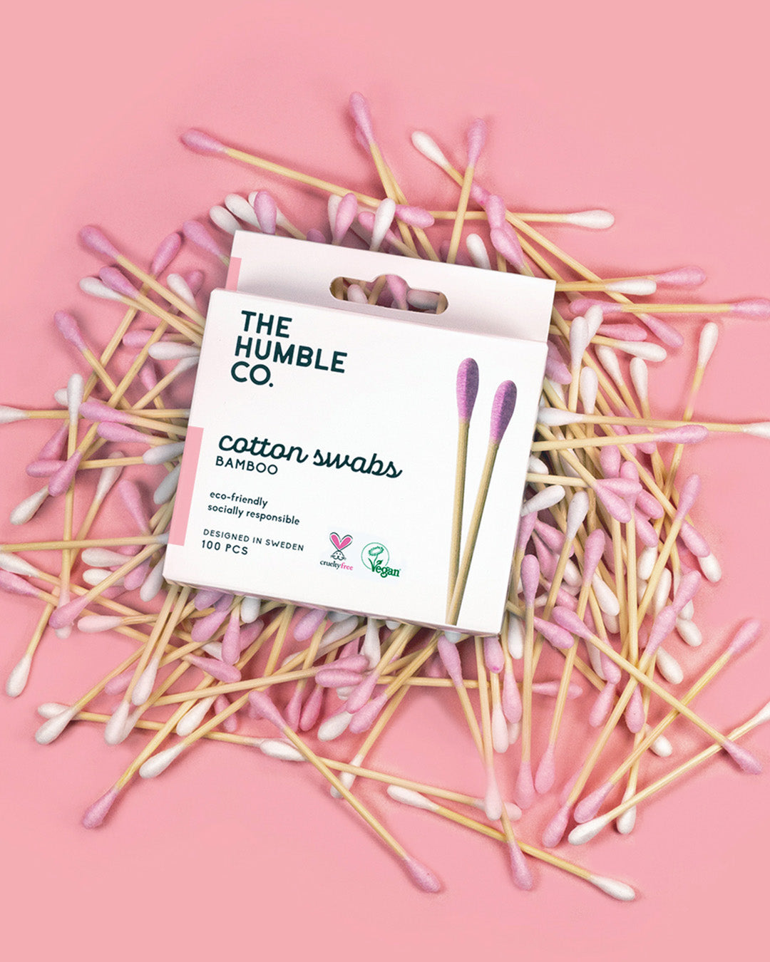 A pack of The Humble Co. cotton swabs with pink tips laid out on a pink background, showcasing the product packaging which emphasizes its biodegradable bamboo handle.