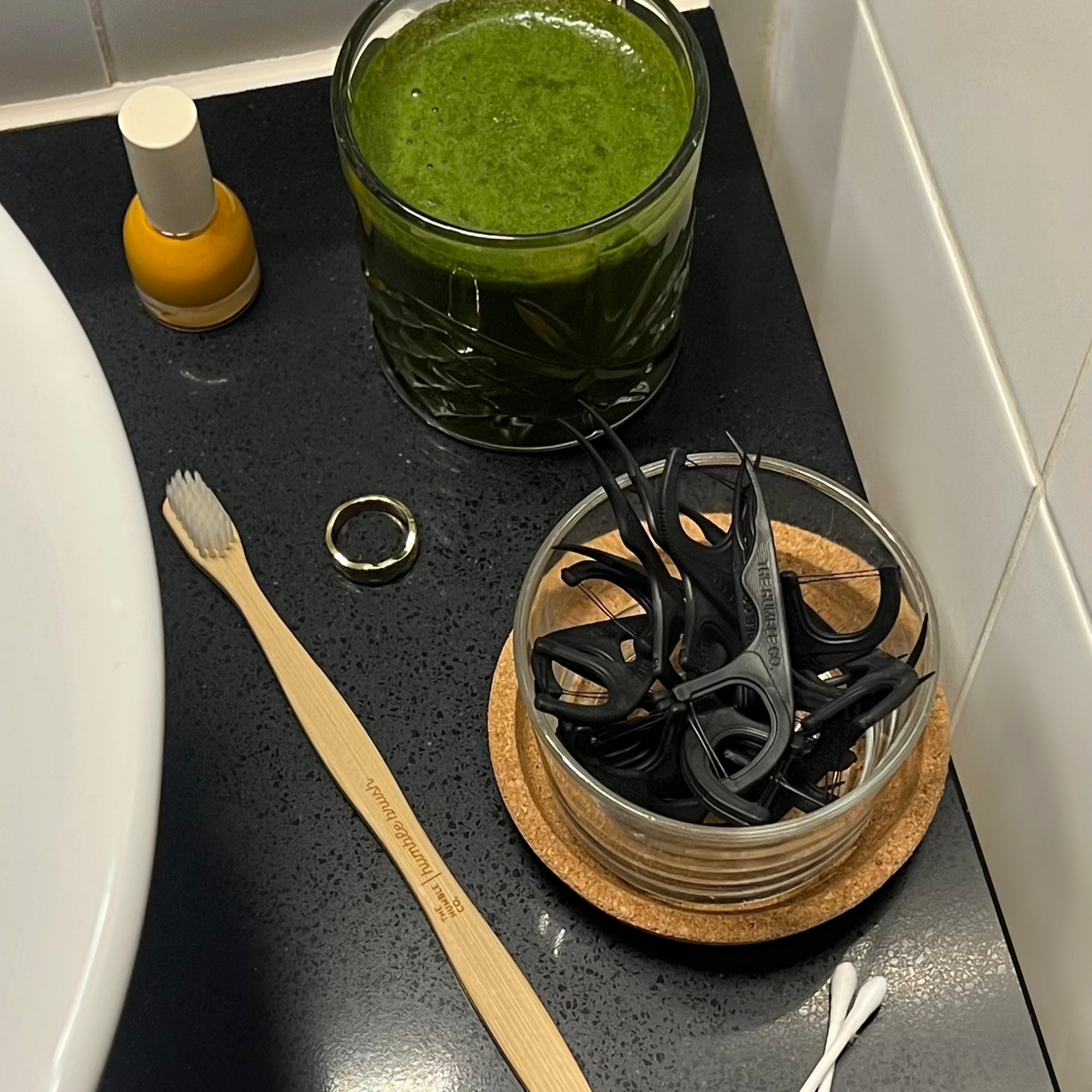 Glass of green juice on a bathroom counter with a toothbrush, scissors, and a ring.