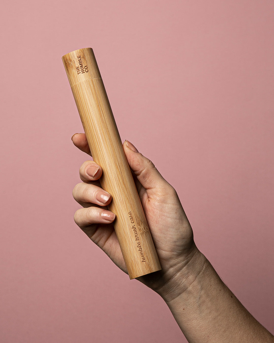 A hand holding a bamboo toothbrush case against a pink background.