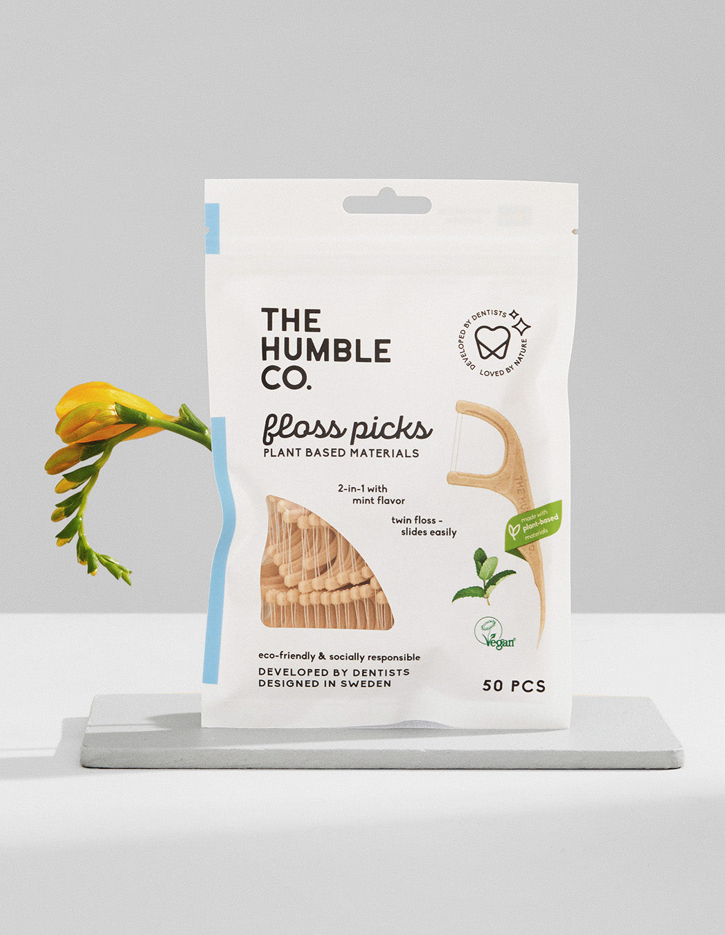 Packaging of 'The Humble Co. Floss Picks - Mint' with 50 pieces of plant-based floss picks displayed in a white and green pouch, with a Freesia flower partially visible to the left.