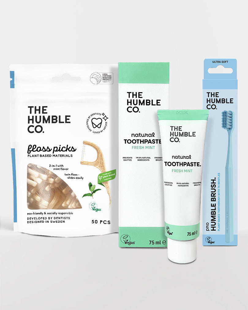 Collection of The Humble Co. dental care products including floss picks, toothpaste, and a toothbrush on a light gray background.