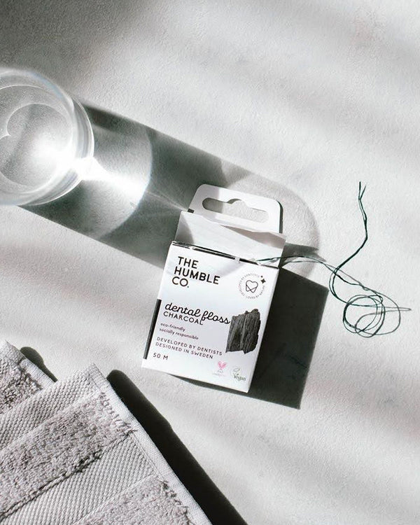 A box of The Humble Co. Dental Floss with charcoal, featuring a sleek design and eco-friendly packaging in a bathroom environment next to a towel and a water glass.